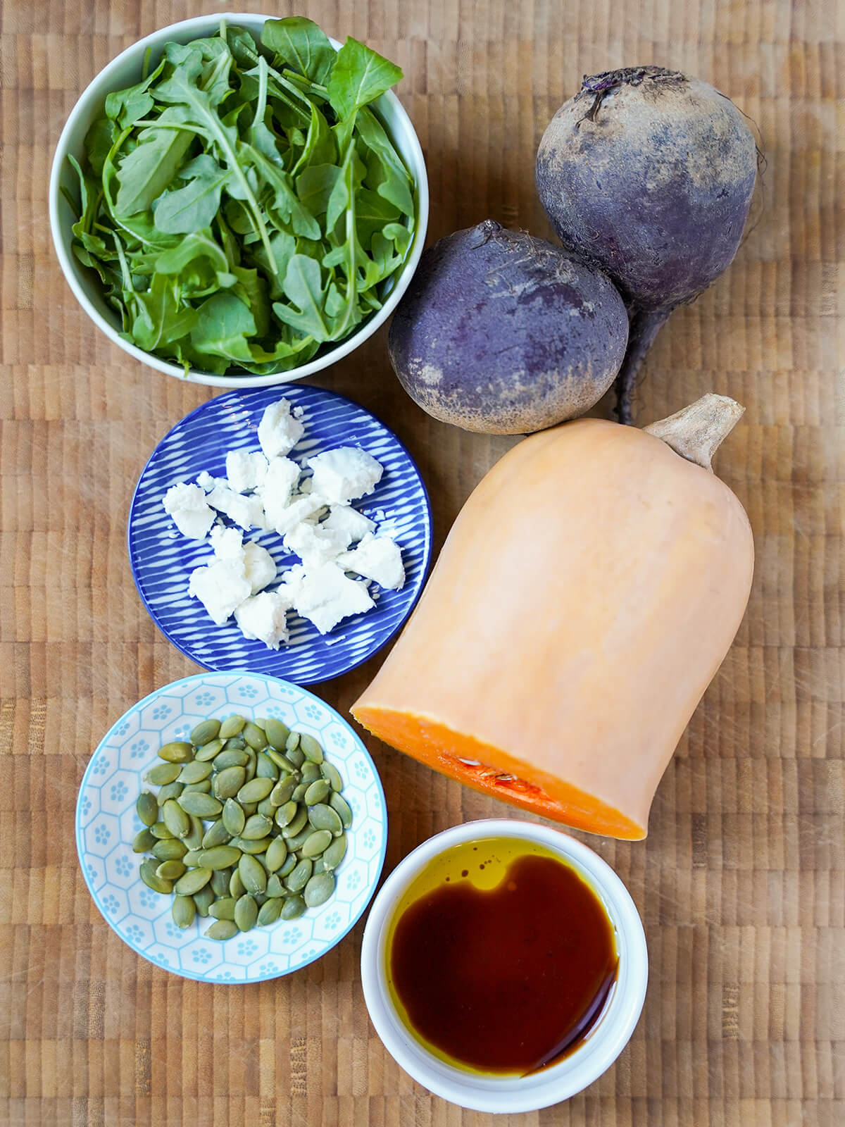 whole beets, part of a butternut squash, bowl of arugula, some pepitas, goat cheese on a dish and dressing ingredients in a bowl.