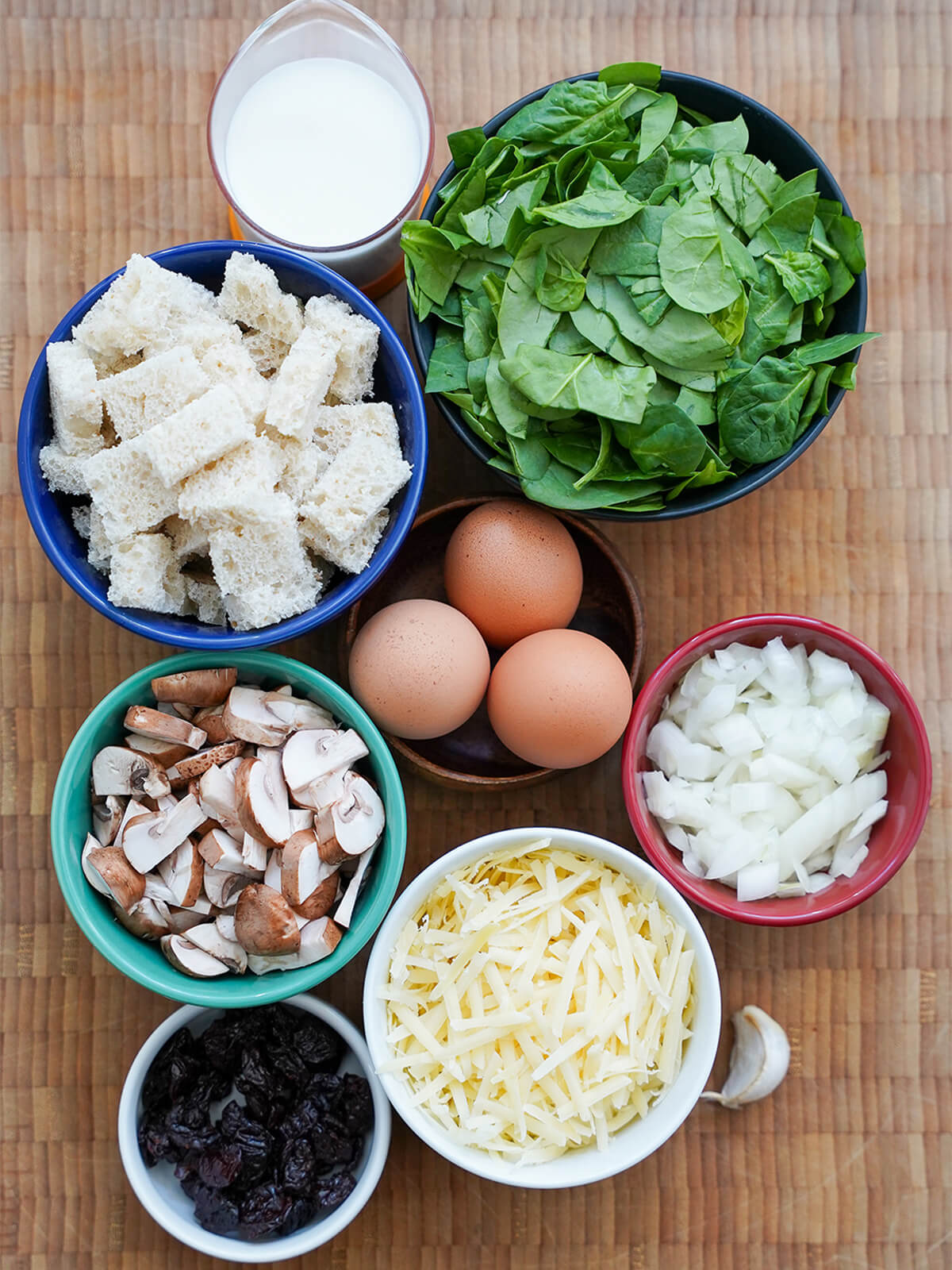 bowls of spinach, chunks of bread, sliced mushroom, onion, grated cheese, dried cherries, eggs and measure of milk.