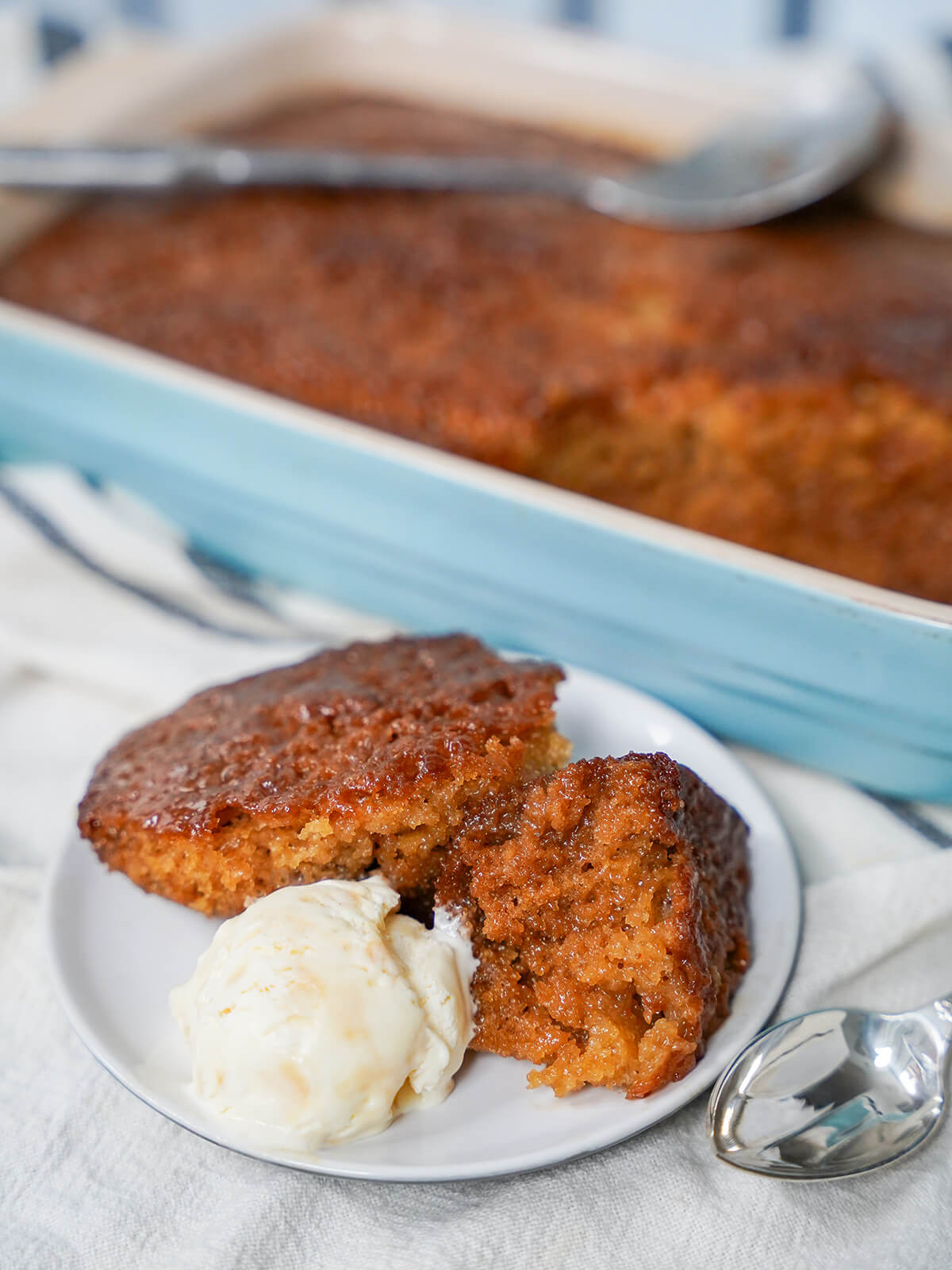 scoops of malva pudding on a plate with a scoop of ice cream with dish and spoon on top of rest of pudding behind.