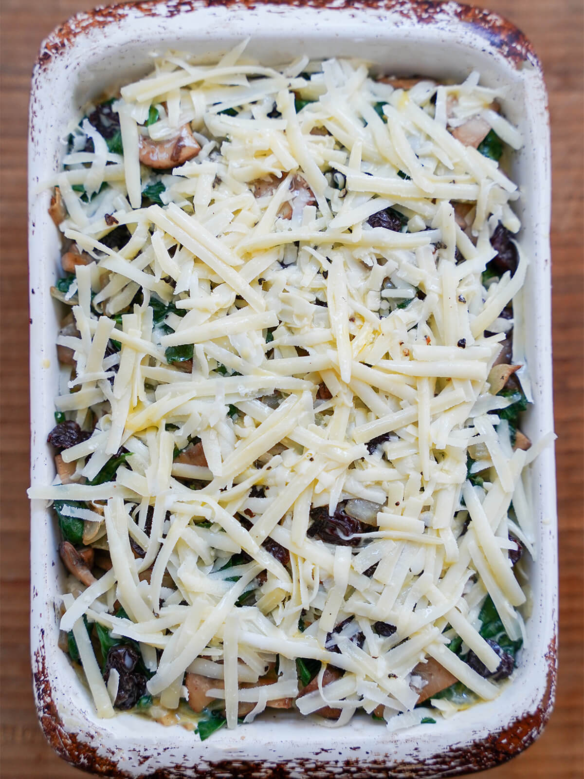 baking dish with layers of strata in it with cheese on top and vegetables partly visible under before cooking.