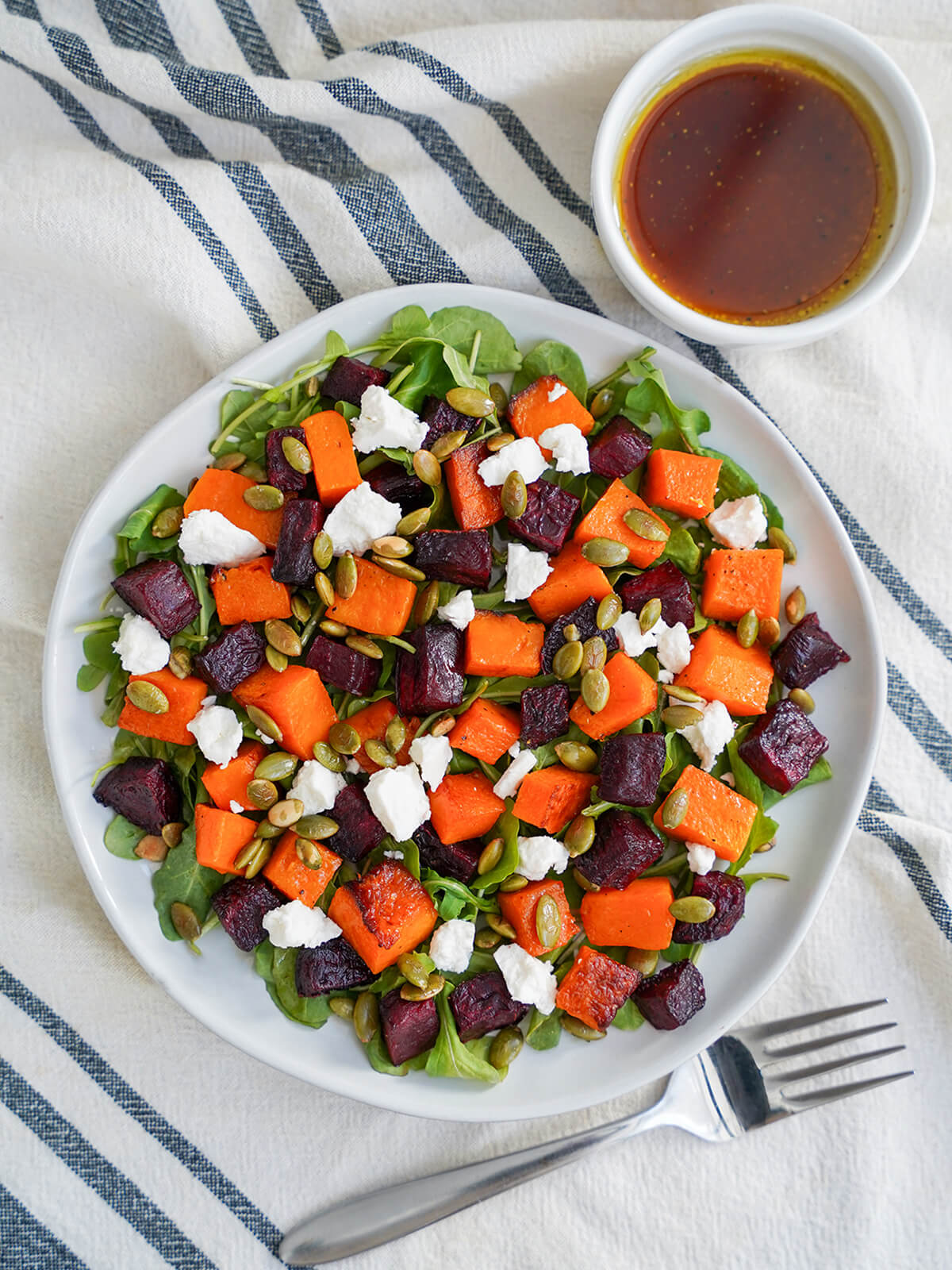 overhead view of plate of roasted beet and butternut squash salad with small bowl of dressing above and fork below plate.