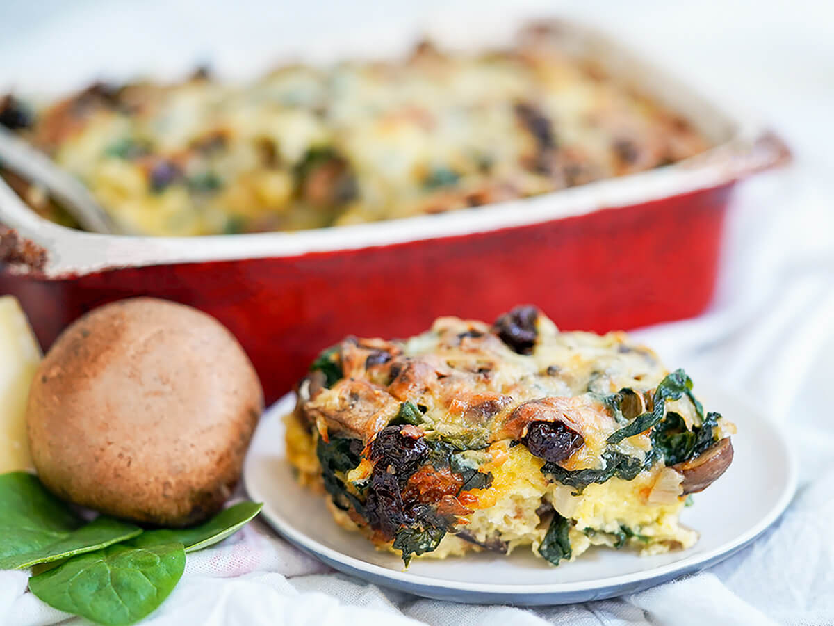 serving of vegetable strata on plate in front of dish with rest, mushroom and some spinach to side.