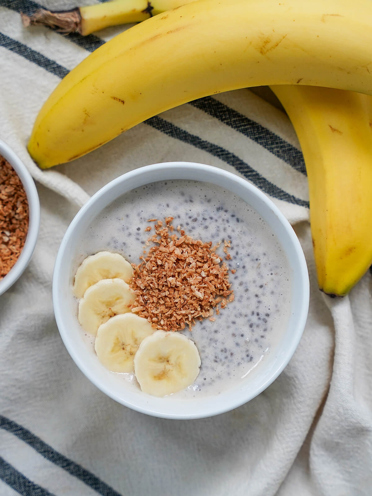 bowl of banana chia pudding with bananas above bowl and toasted coconut and banana slice garnishes in bowl.