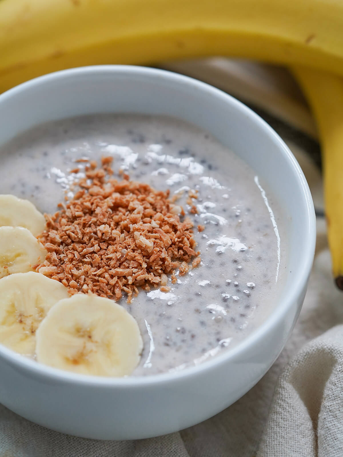 part side view of bowl of banana chia pudding close in with bananas behind.