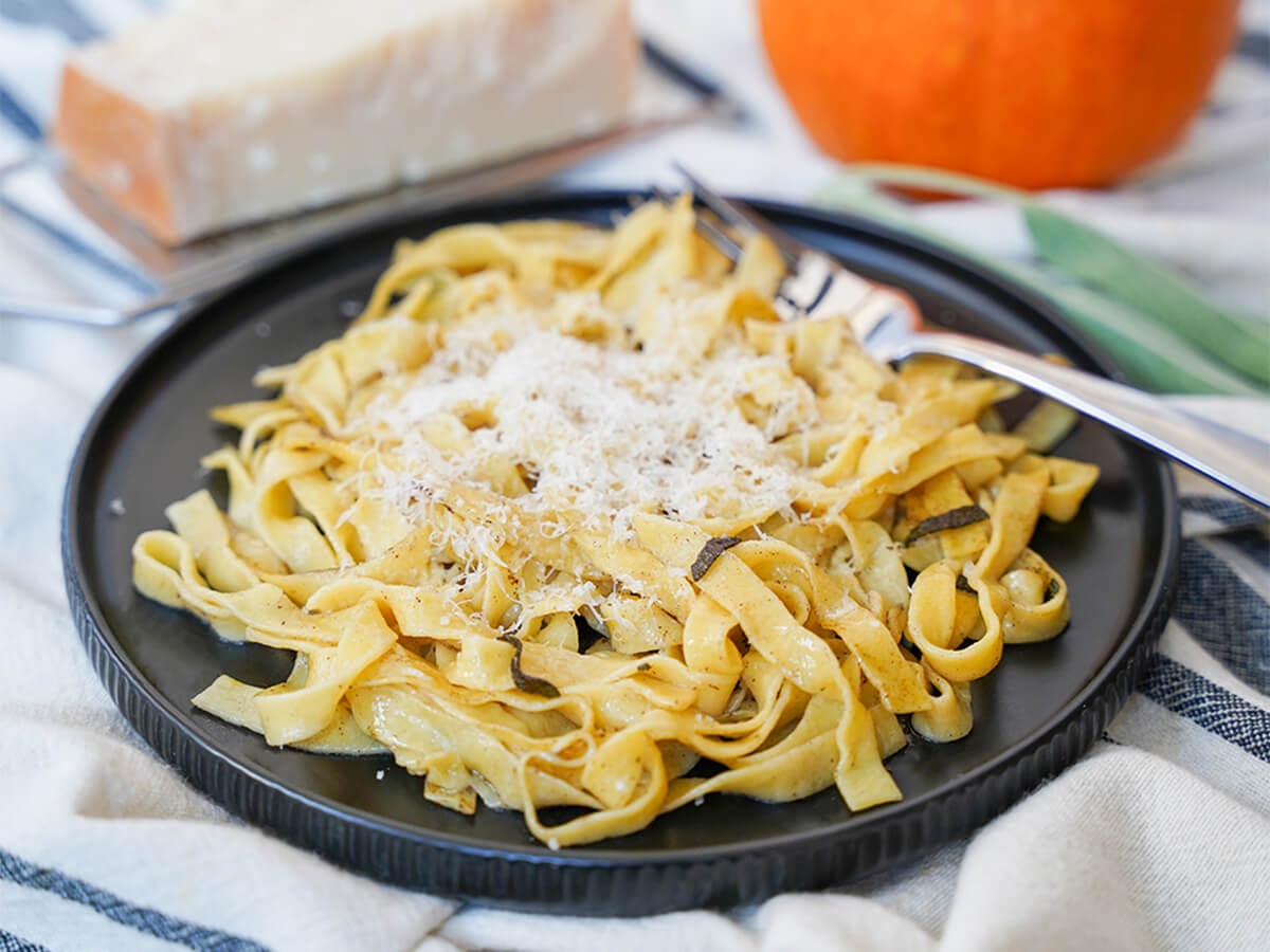 This pumpkin pasta is a delicious way to enjoy a touch of fall in homemade pasta with real pumpkin in the dough. If you're a pasta fan, you'll be blown away at how good it can taste. plate of fresh pumpkin pasta with block of cheese and part pumpkin visible blurred behind.