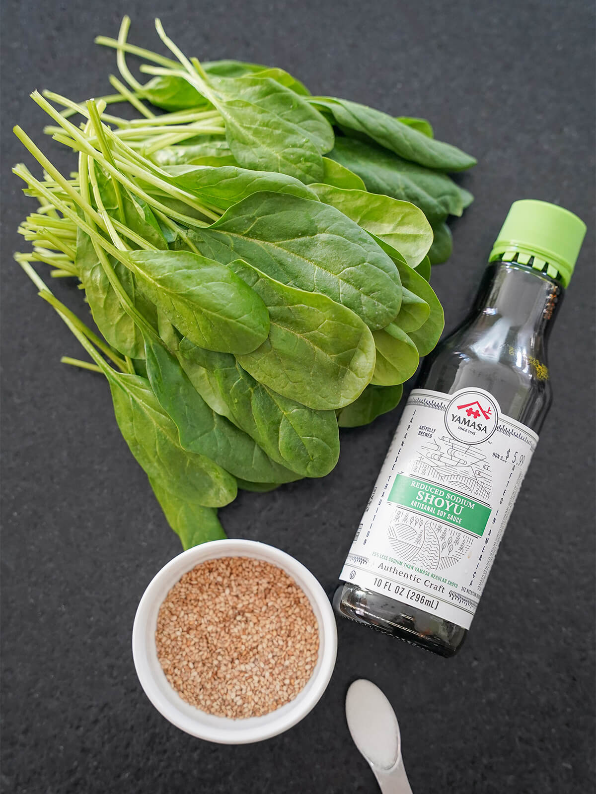 stack of spinach leaves, bottle of soy sauce, bowl of sesame seeds and spoon of sugar.