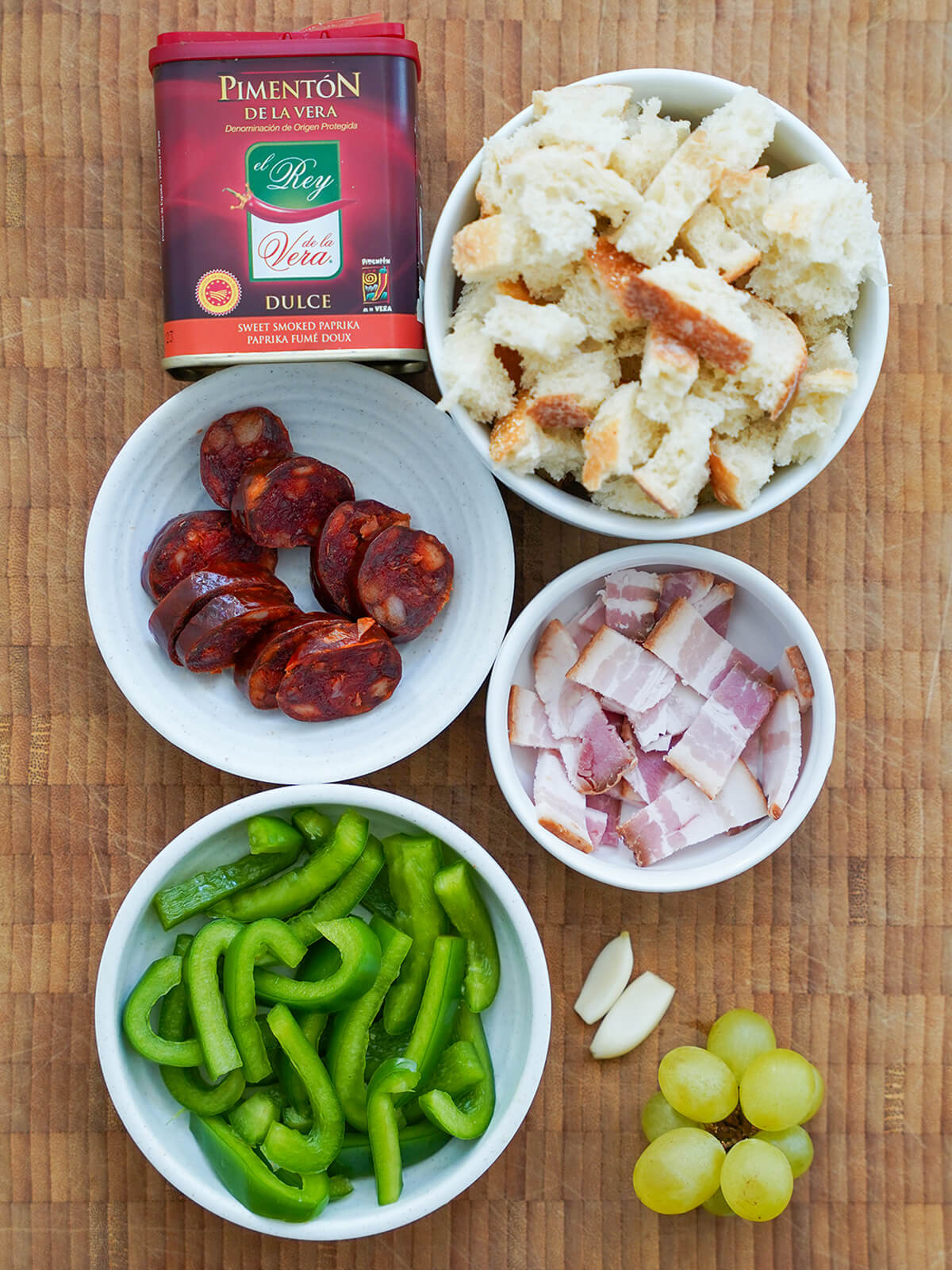 bowls with chunks of bread, chorizo and bacon slices, slices green pepper with garlic and grapes to side.