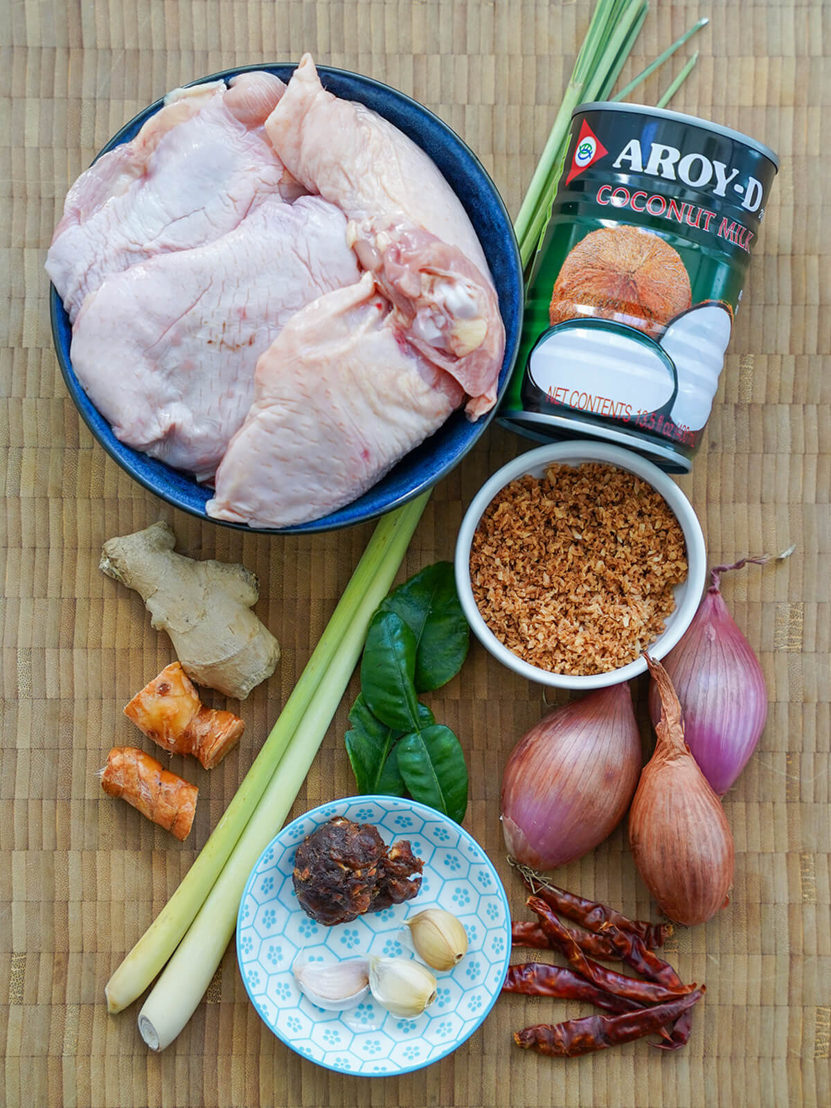 bowl of chicken thighs, can of coconut milk, small dishes with toasted coconut, garlic, knob of tamarind with shallots, chilis, ginger, lemongrass and lime leaves around.