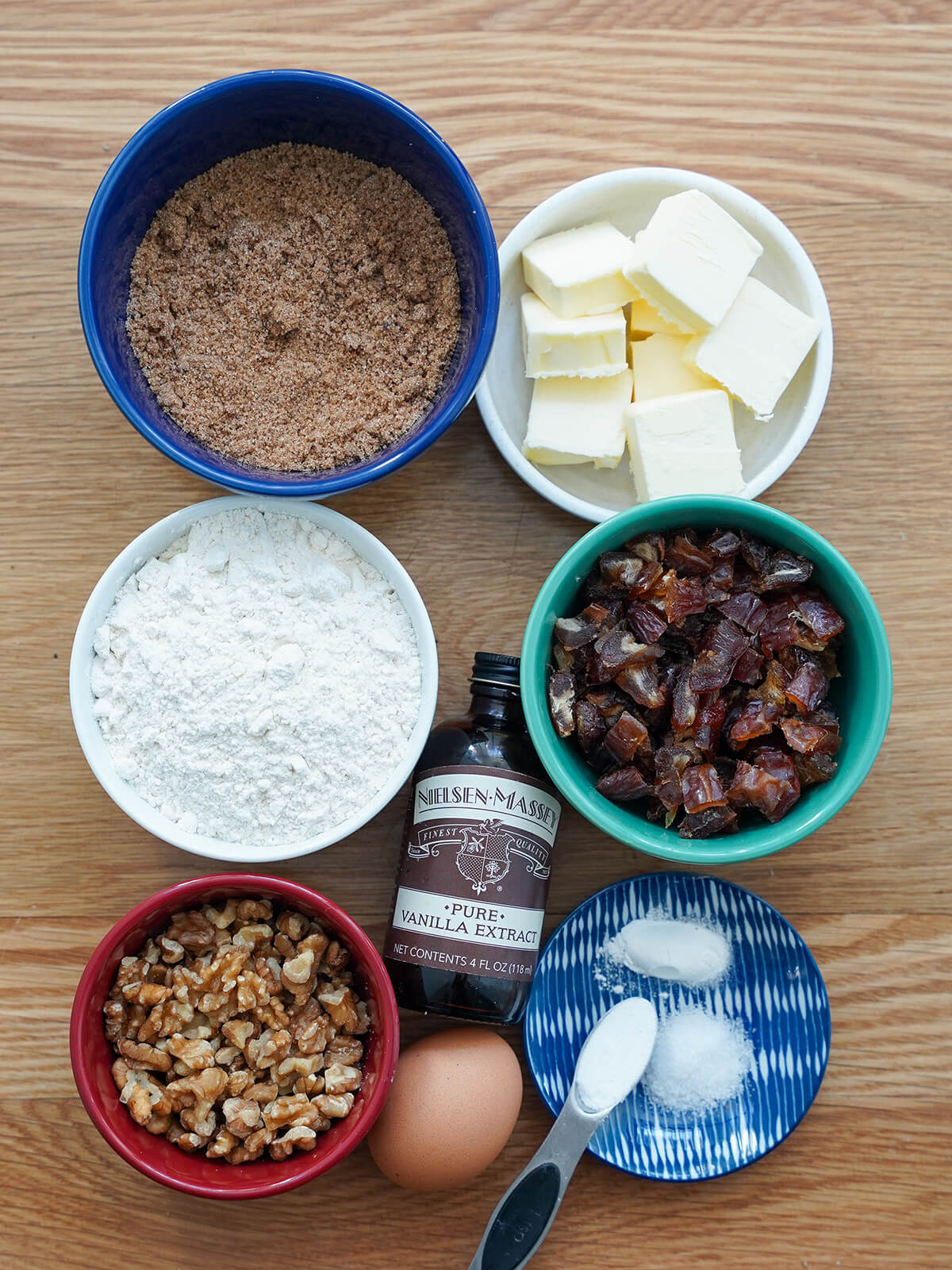 bowls of brown sugar, butter, flour, chopped dates, chopped walnuts with egg, vanilla in between and raising agents on small plate.