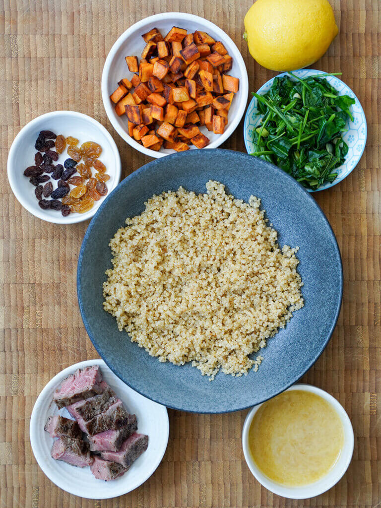 bowl of quinoa with small dishes of sweet potato cubes, sauteed spinach, steak, raisins and dressing around it.