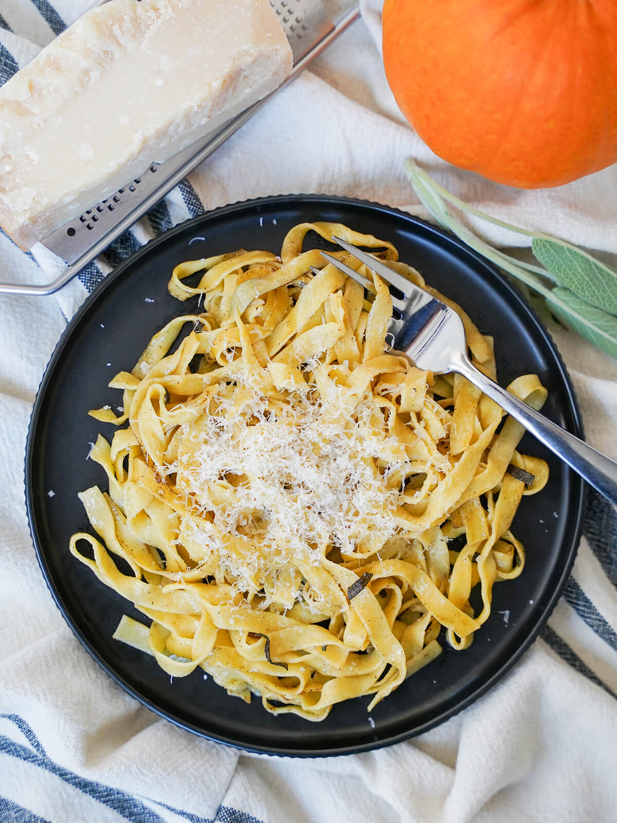 This pumpkin pasta is a delicious way to enjoy a touch of fall in homemade pasta with real pumpkin in the dough. With a subtle savory flavor, and everything that makes homemade pasta so good. overhead view of a plate of fresh pumpkin pasta with block of parmesan on grater above and pumpkin and sage to side.