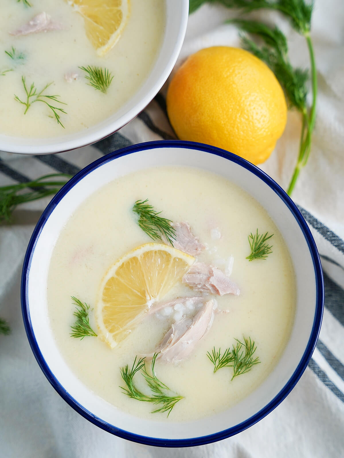 overhead view of a bowl of avgolemono soup with lemon and dill above and part of a second bowl in corner.