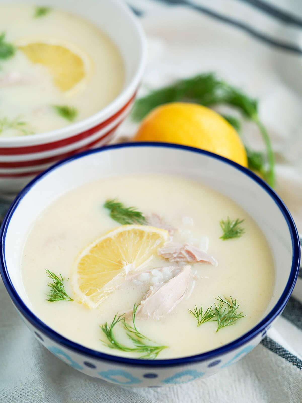 sde view of a bowl of avgolemono soup with top of lemon and bunch of dill blurred behind.