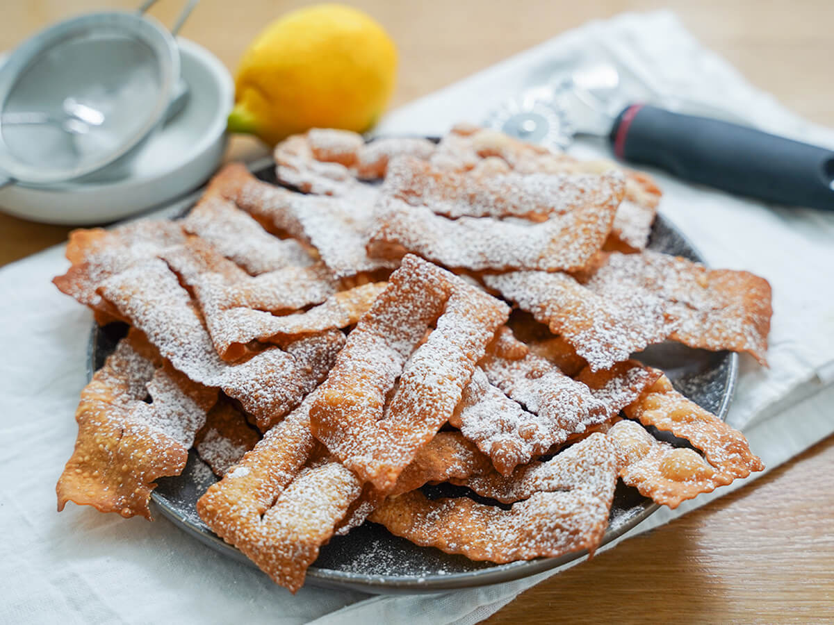plate of chiacchiere, fried pastries, from side with lemon and ravioli cutter blurred behind.