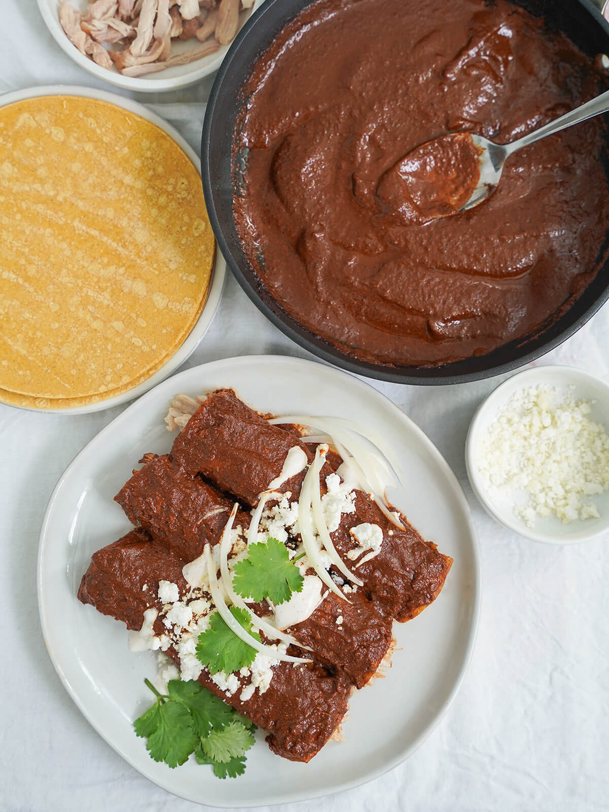 plate of enmoladas with mole sauce and corn tortillas to side. overhead view of three enmoladas on a plate with skillet of mole and plate of corn tortillas above.