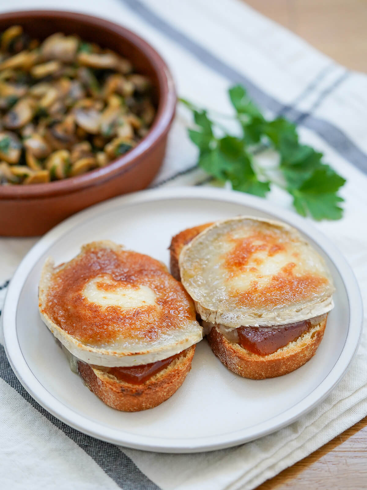 two pieces of goat cheese toast with dish of mushrooms and sprig of parsley blurred in background.