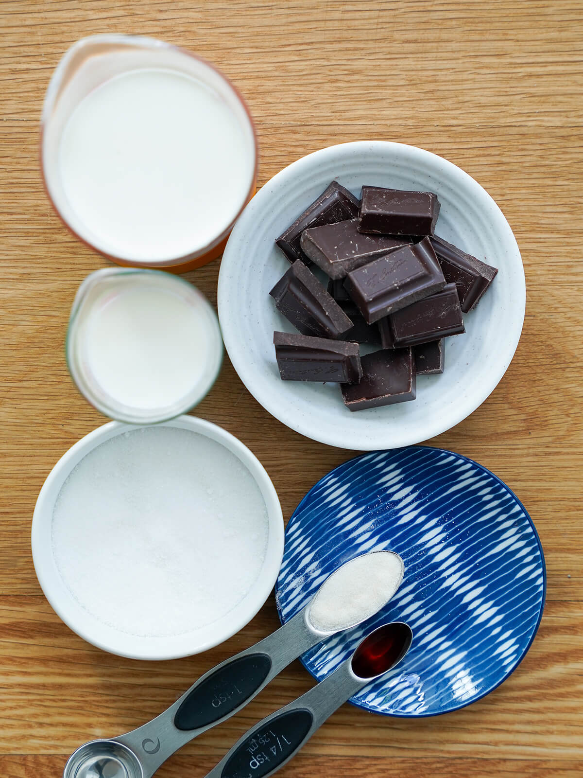 dishes with chunks of chocolate, sugar, spoons of vanilla and gelatin and measures with cream and milk.