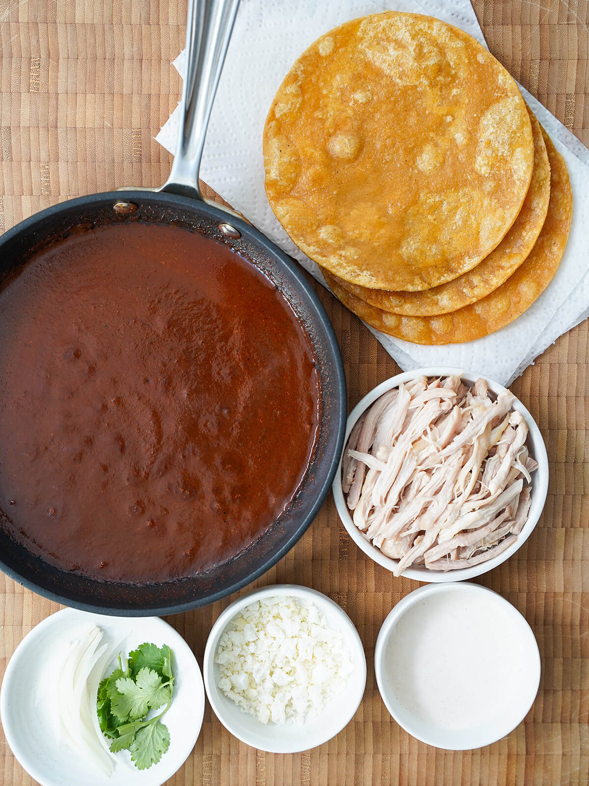 components for enmoladas skillet of mole sauce, fried corn tortillas, dish of shredded chicken, dishes with crema, cheese and onion slices.