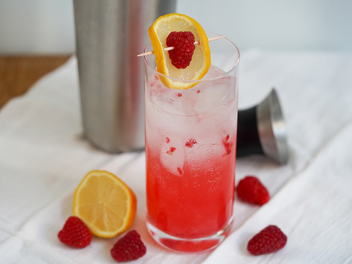 raspberry collins cocktail with raspberries and lemon to side.