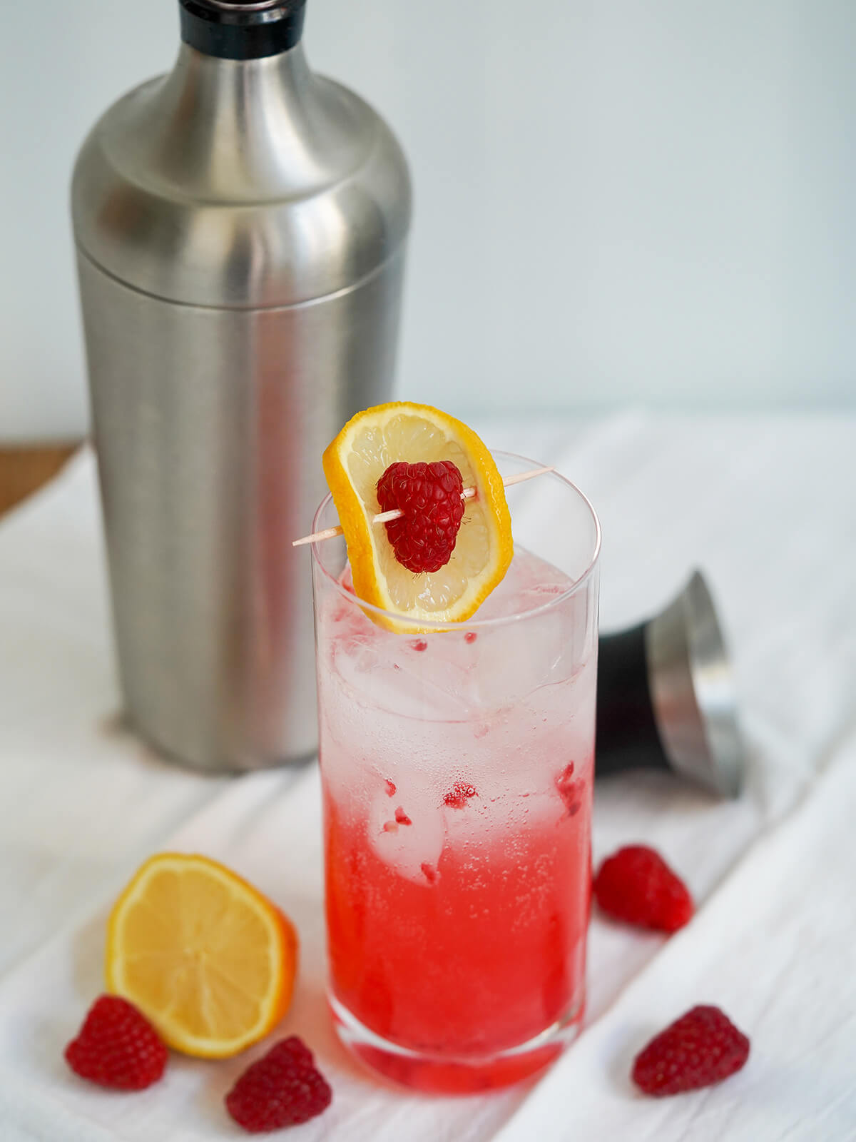 glass of raspberry collins cocktail with lemon raspberry garnish and cocktail shaker behind.