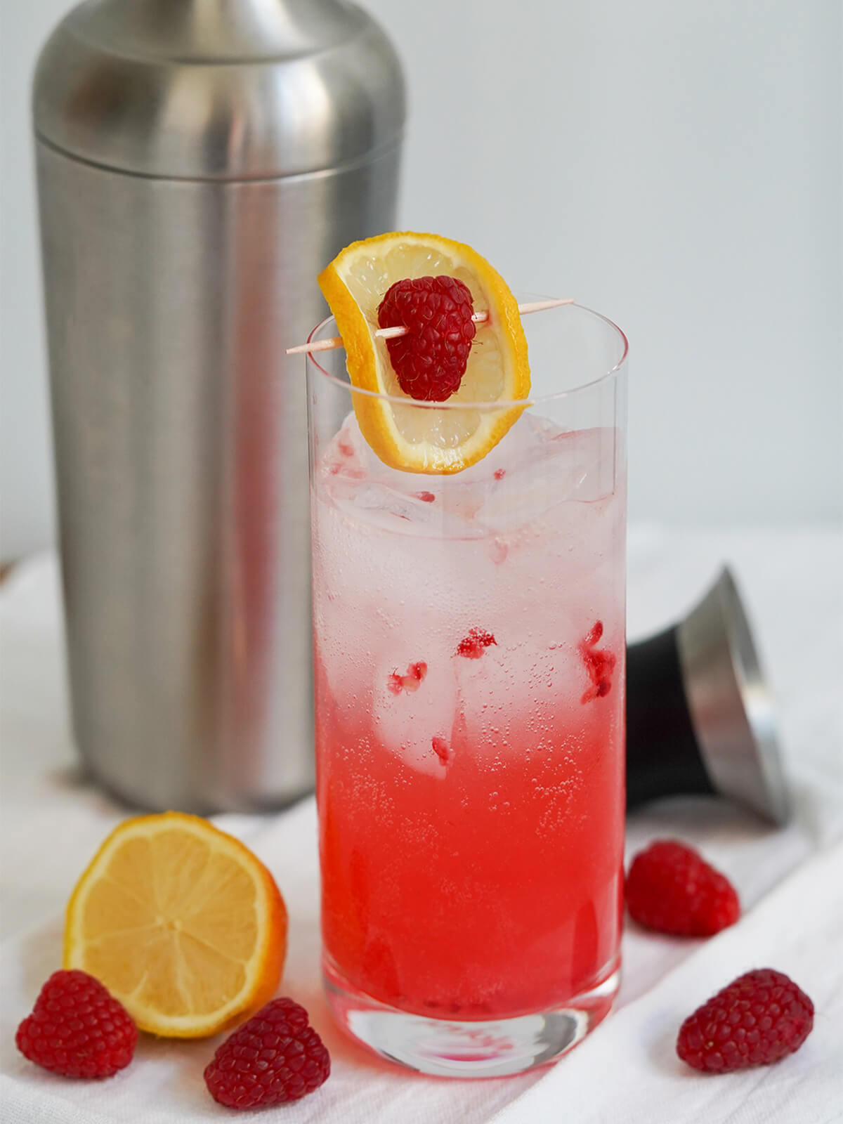 glass of raspberry collins cocktail with cocktail shaker behind and fruit to side.