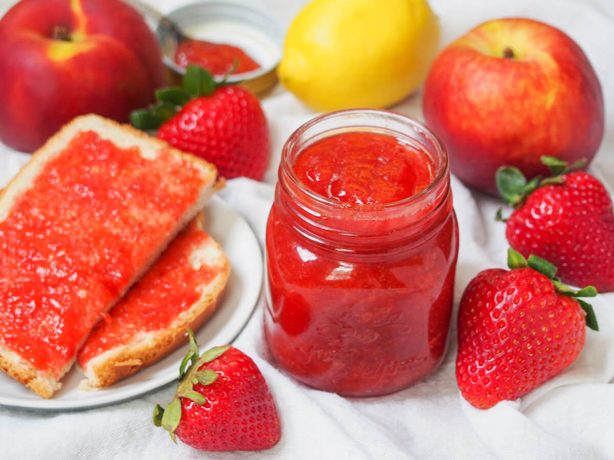 small jar of strawberry peach jam with pieces of toast topped with jam to side and strawberries around and peaches and lemon behind.