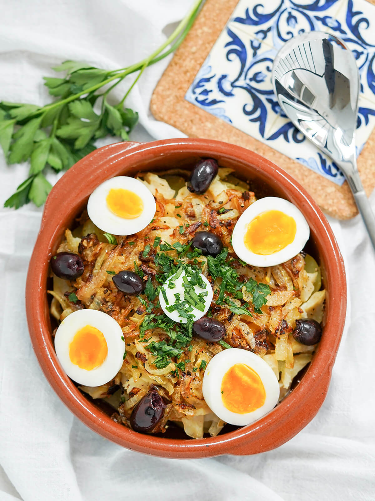 overhead view of dish oof Bacalhau à Gomes de Sá, Portuguese salt cod with spoon to side on tiled mat.