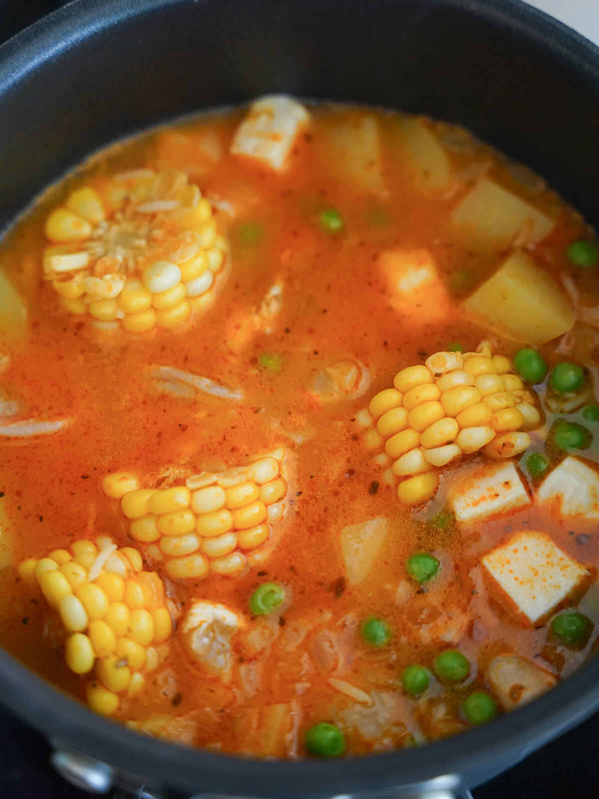 chunks of corn cob, potato, cheese, peas showing on top of soup in pot.