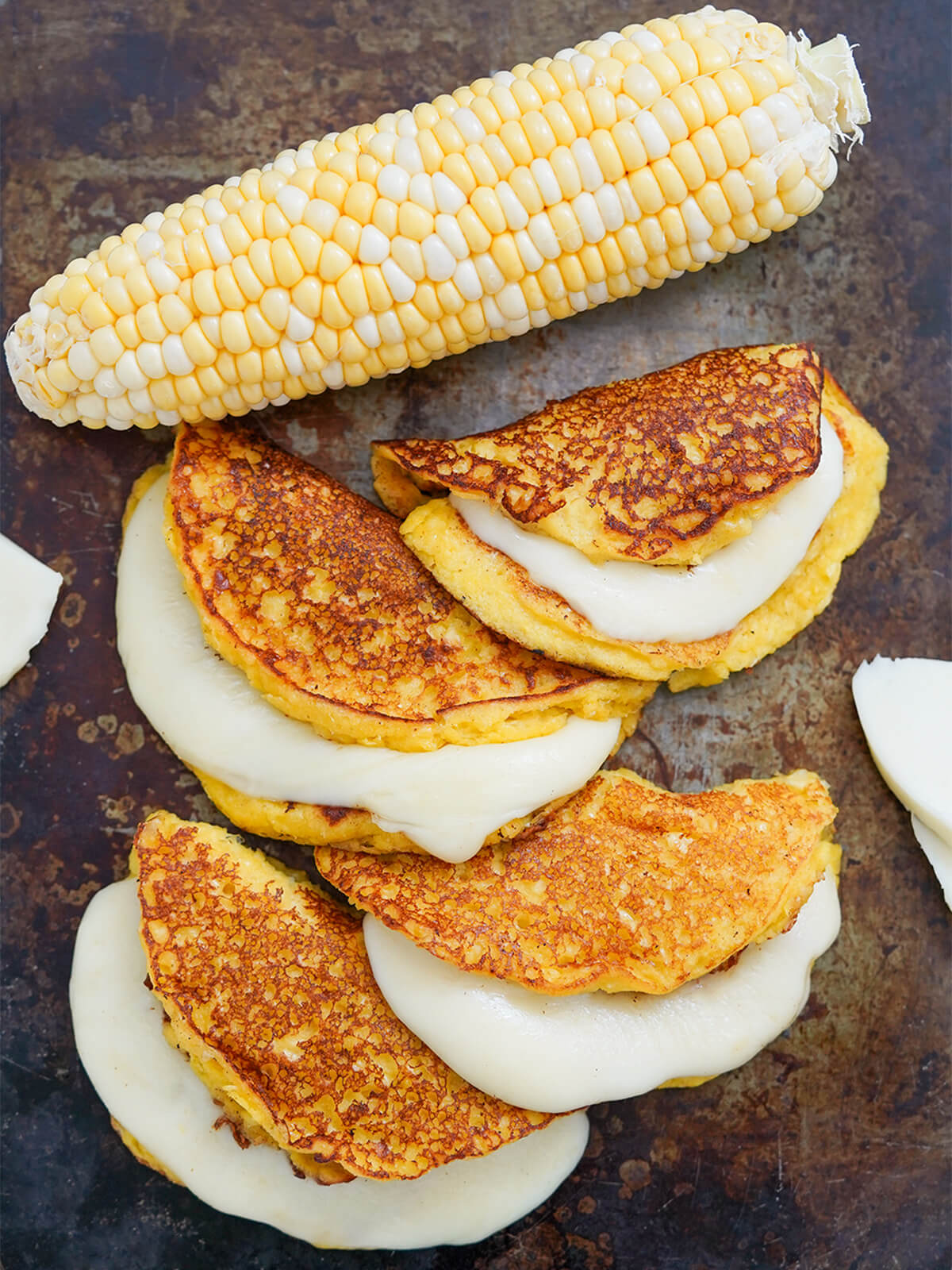 overhead view of four cachapas, Venezuelan corn pancakes with ear of corn above.