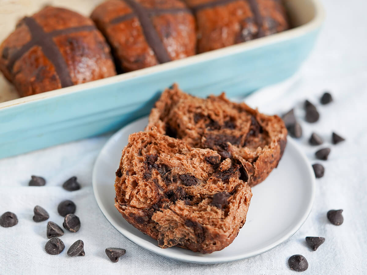 chocolate hot cross buns in dish behind one split open on plate with chocolate chips around.