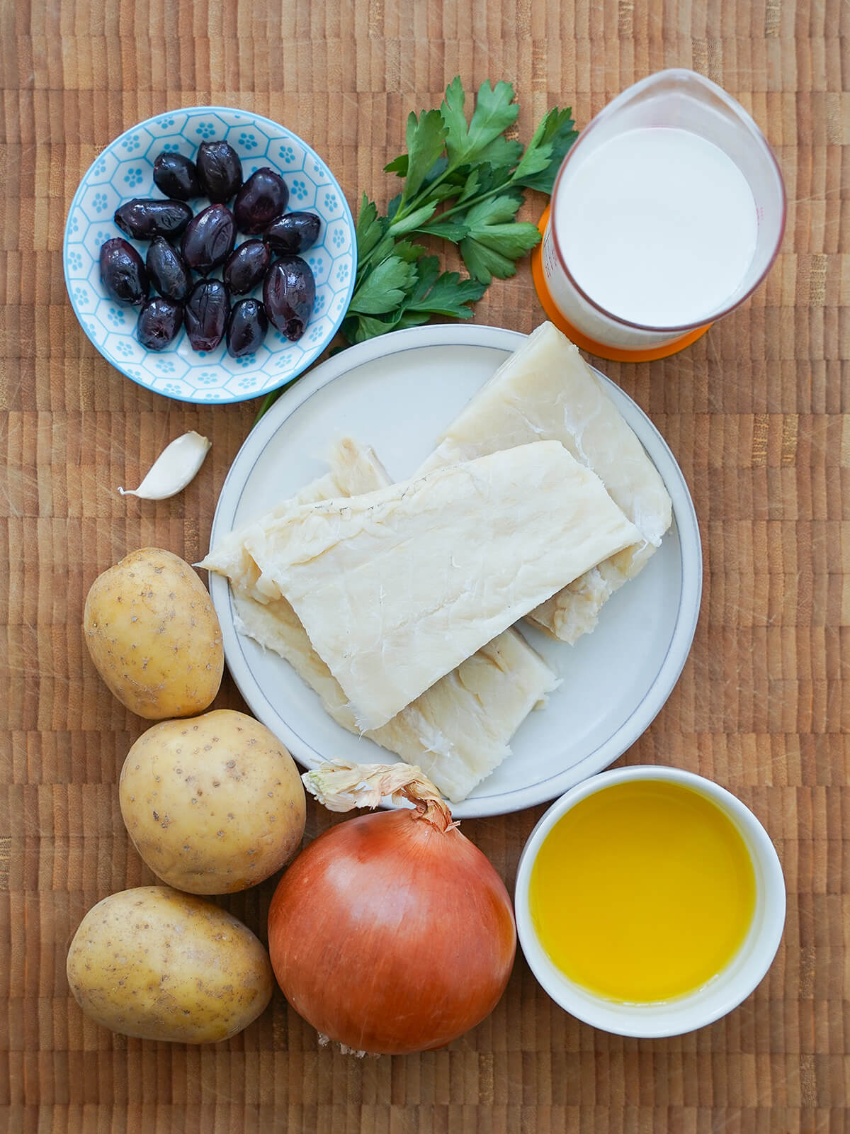 plate of salt cod with dishes with olives, olive oil, milk to side and onion and potatoes below.