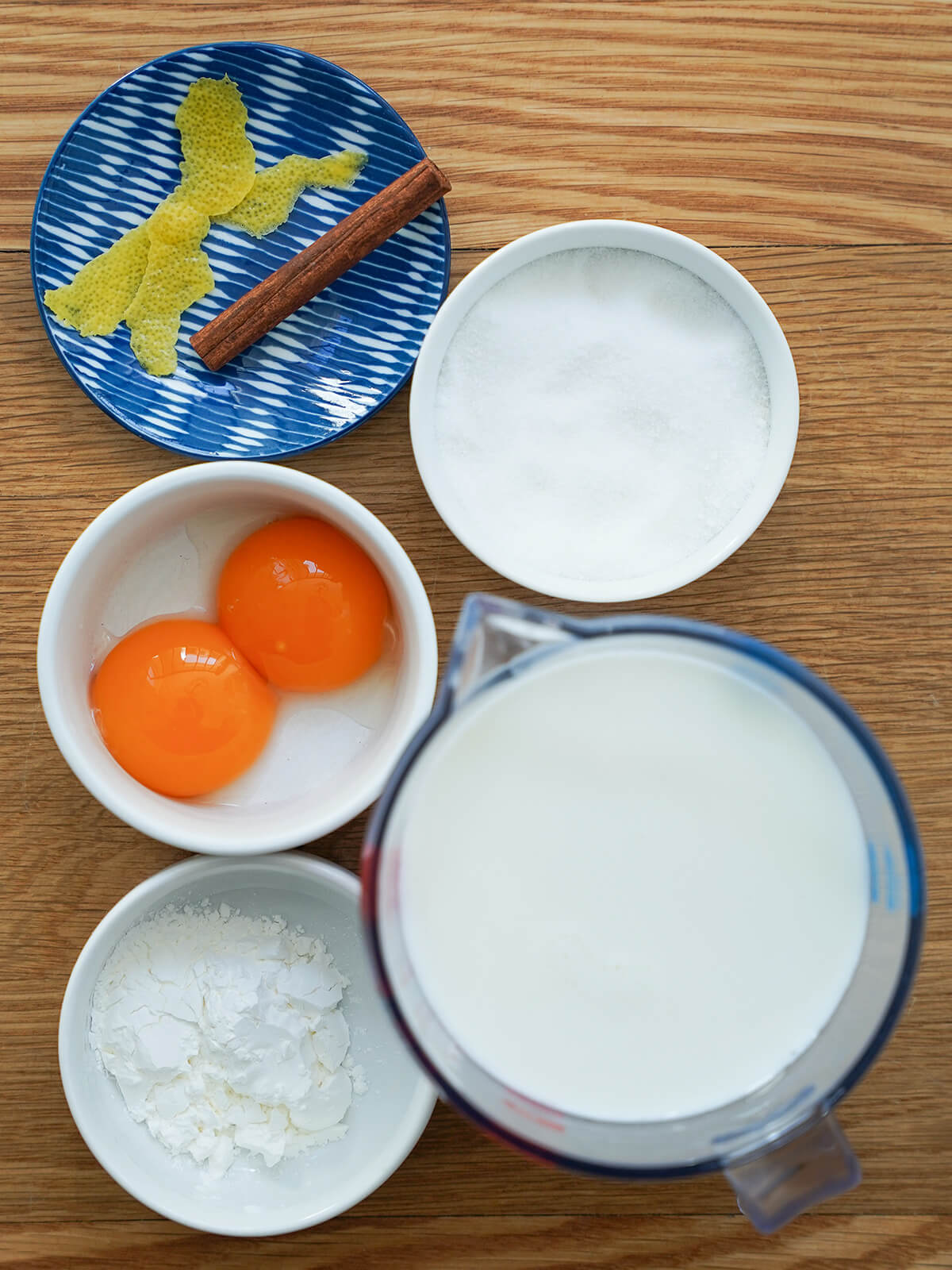 measure of milk, bowls with egg yolks, sugar, corn starch and small plate with strips of lemon peel and cinnamon stick.