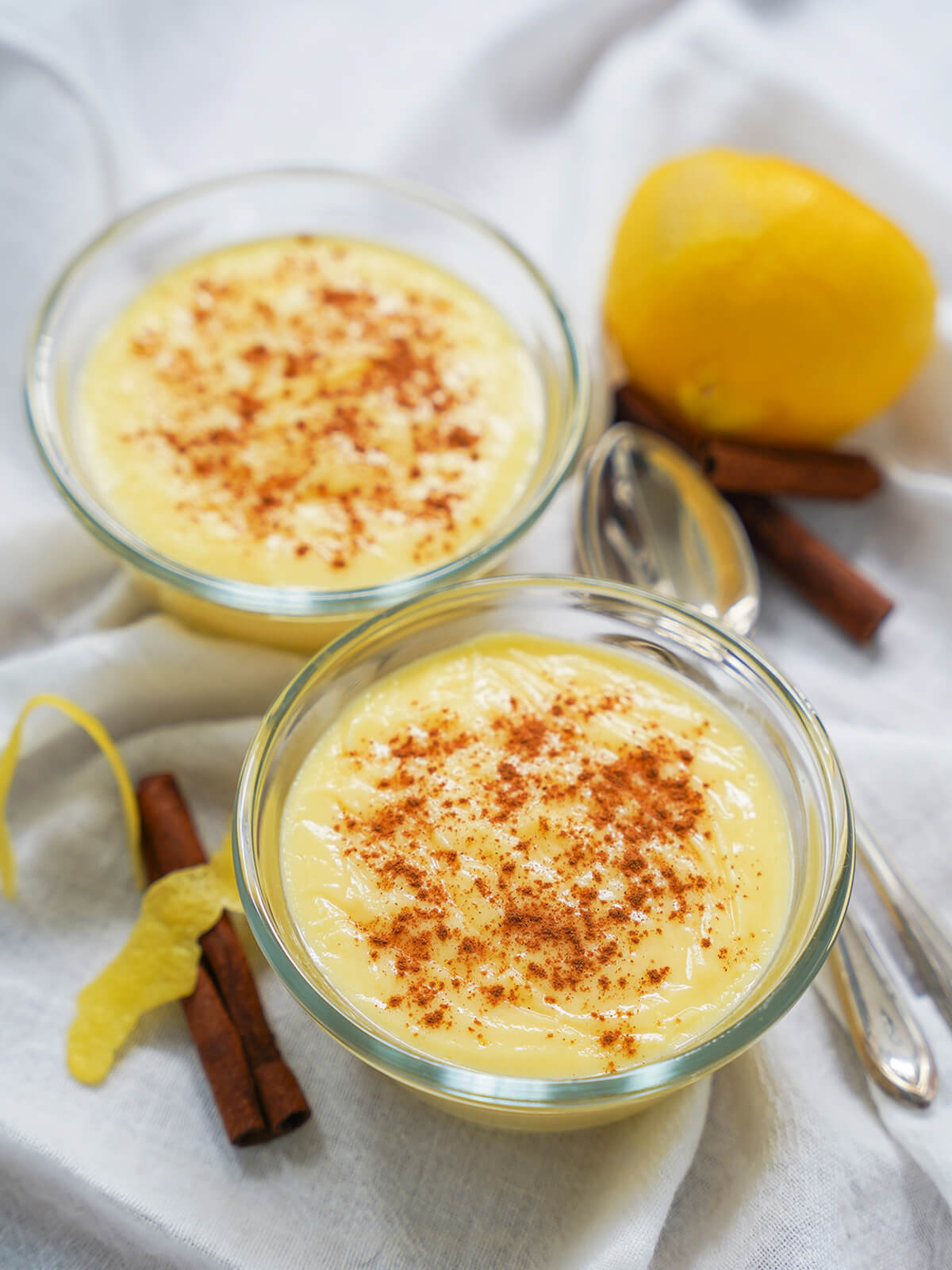 two dishes of natillas, Spanish custard, with part lemon behind, cinnamon sticks behind and to side.