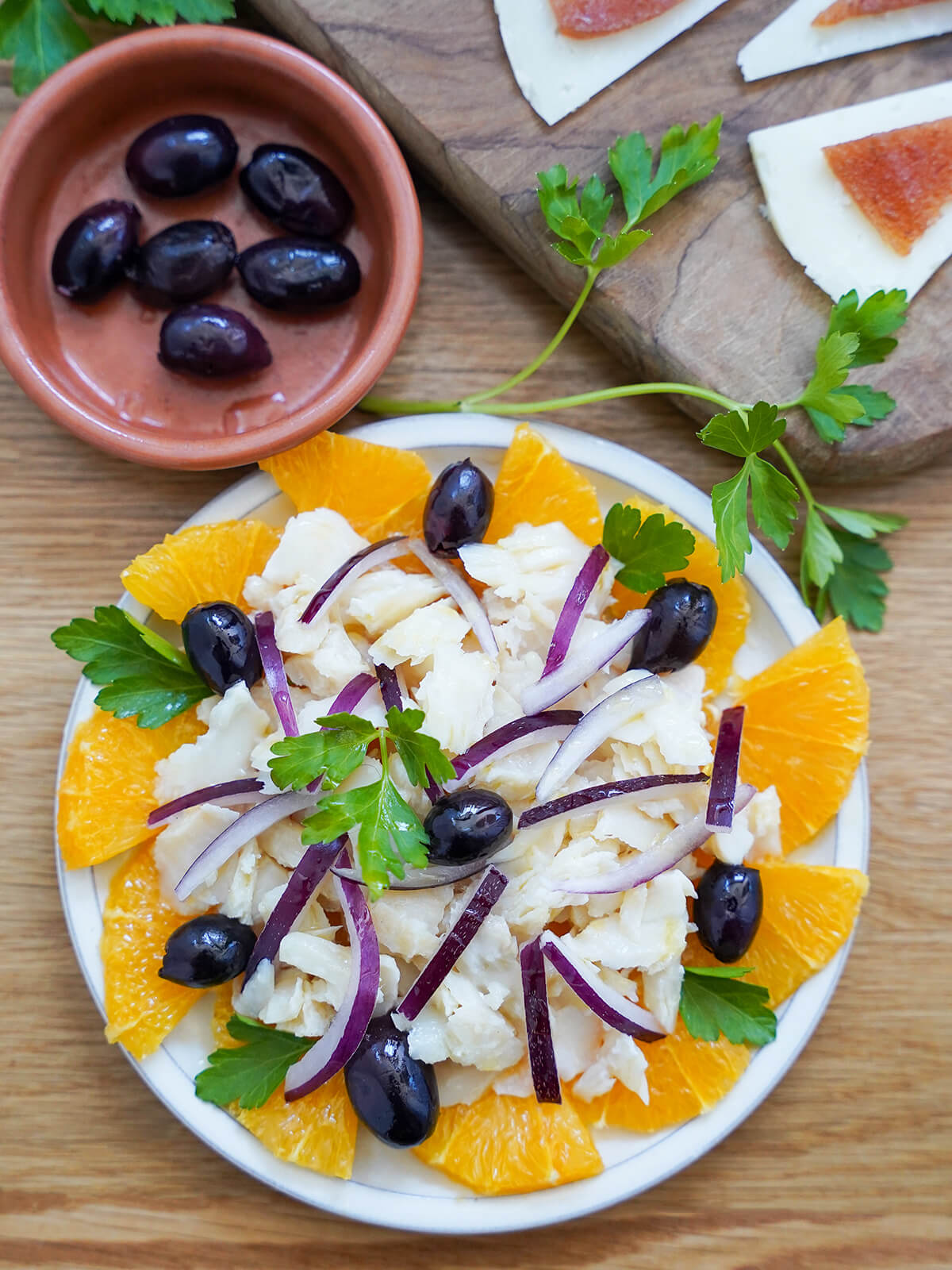 overhead view of plate of remojon, salt cod orange salad with dish with olives and pieces of cheese with membrillo above.