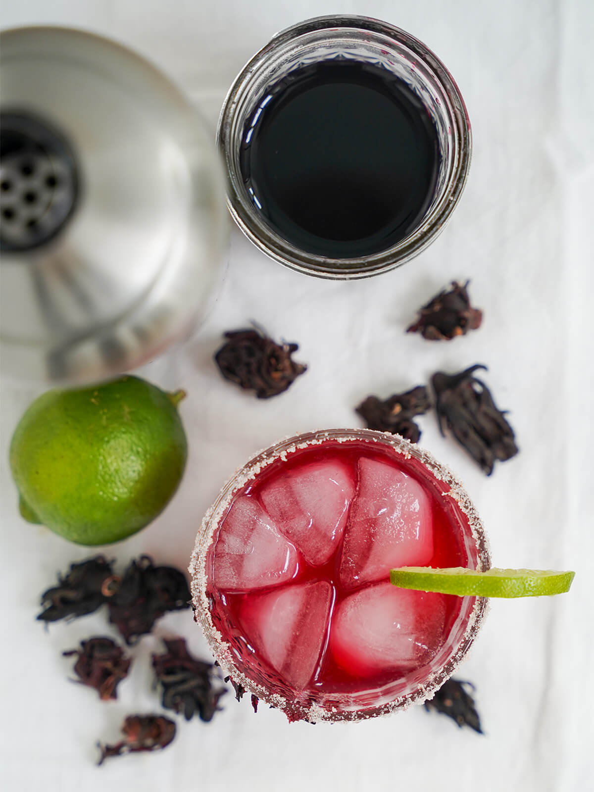 overhead view of hibiscus margarita with ice cubes in top and lime and hibiscus flowers to side of glass.
