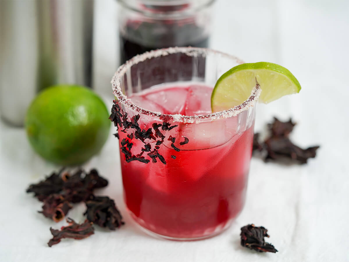 glass of hibiscus margarita with lime and hibiscus flowers to side of glass.