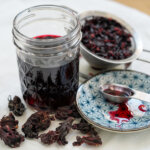 jar of hibiscus syrup with spoon and a little syrup on plate to side, drained flowers behind.