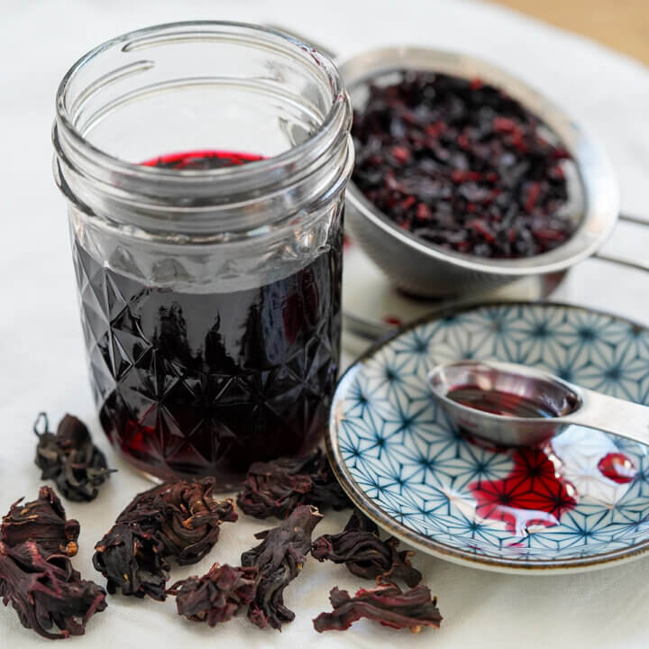 jar of hibiscus syrup with spoon and a little syrup on plate to side, drained flowers behind.