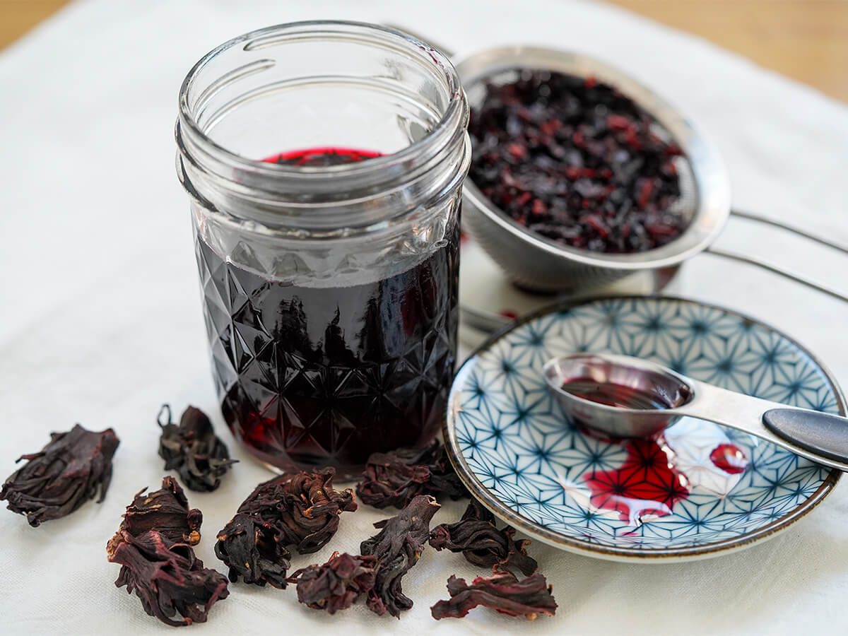 jar of hibiscus syrup with small plate to side with drops of syrup and spoon on it, flowers around jar.