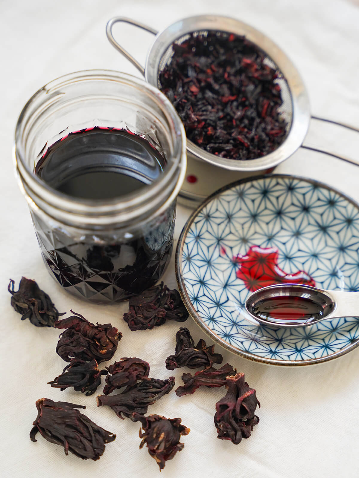 view looking into jar of dark colored hibiscus syrup with some on spoon on plate to side a brighter red.