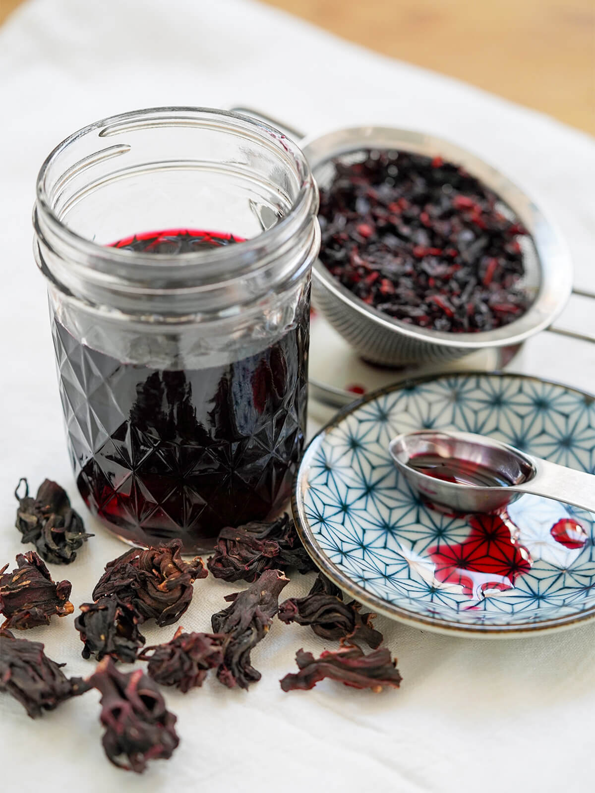 jar of hibiscus syrup with strained flowers behind, whole flowers in front and drops of syrup on plate to side.