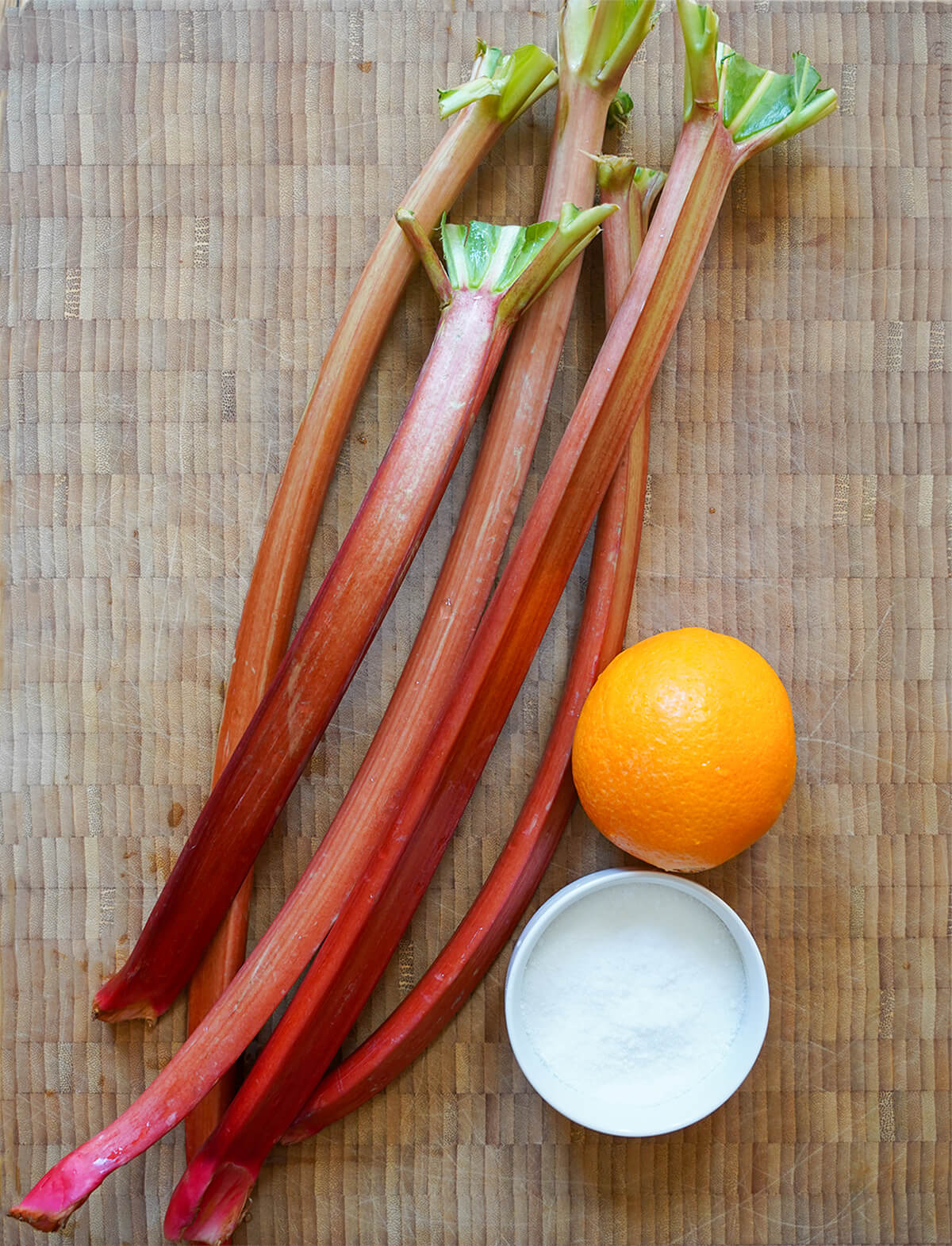 stems of rhubarb, an orange oand bowl of sugar on wooden board.