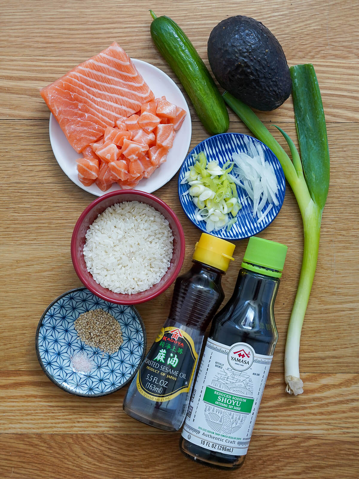 plate with salmon, small plates with sliced onion and sesame seeds, cucumber, avocado above, bowl of rice and bottles of soy and sesame oil.