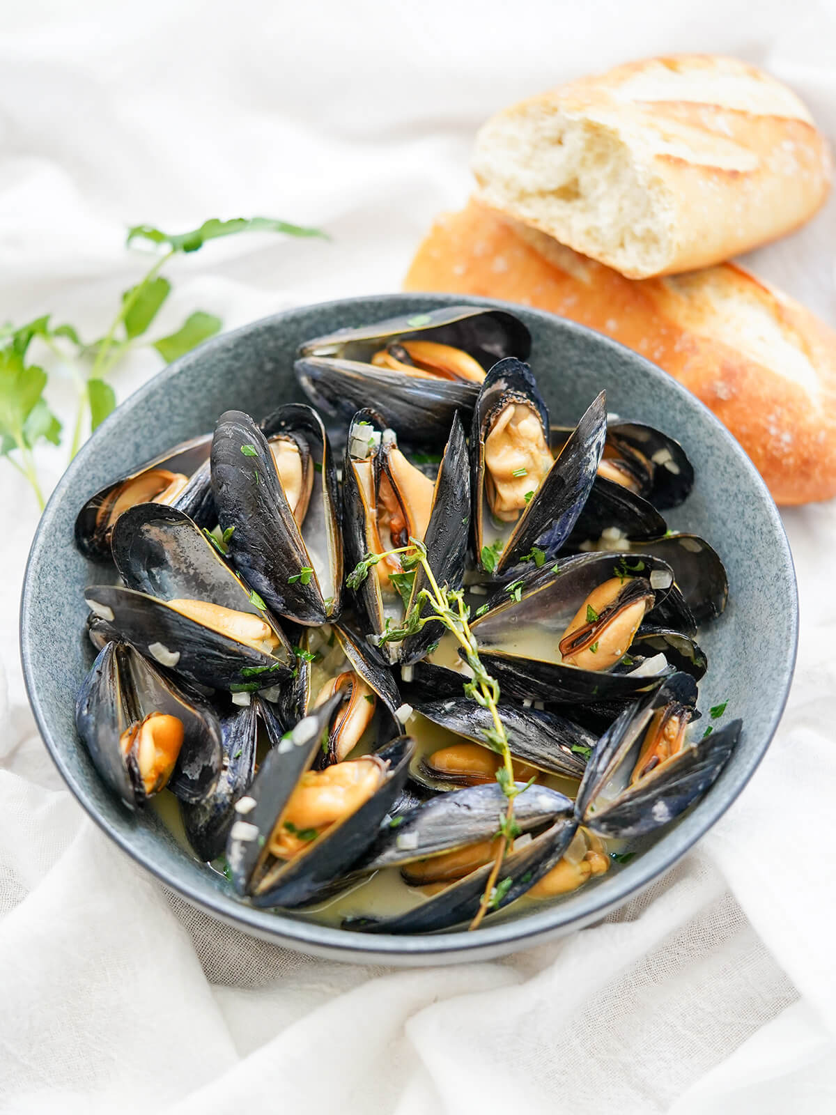 Mussels in cream is a wonderfully easy seafood dish of mussels in a light, creamy and flavorful broth. It's quick, relatively inexpensive and delicious. side view of mussels in cream with split baguette and parsley behind bowl.