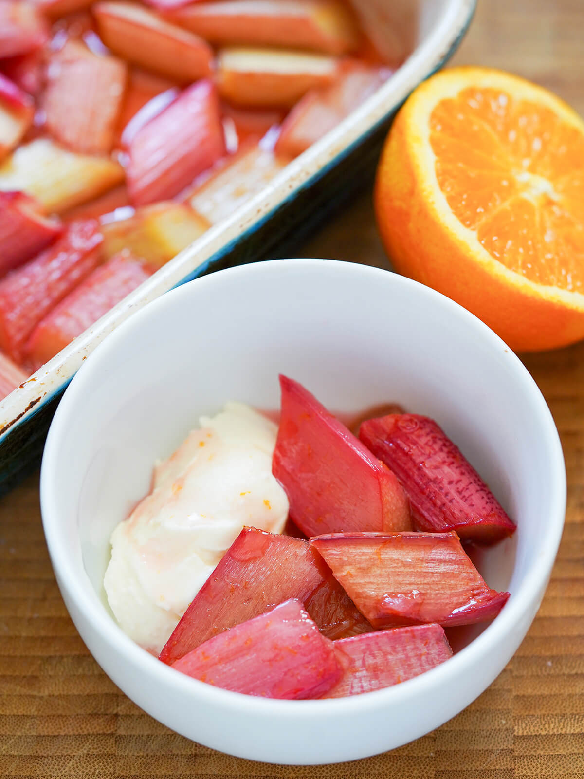 bowl of roasted rhubarb with baking dish with more and half orange behind.