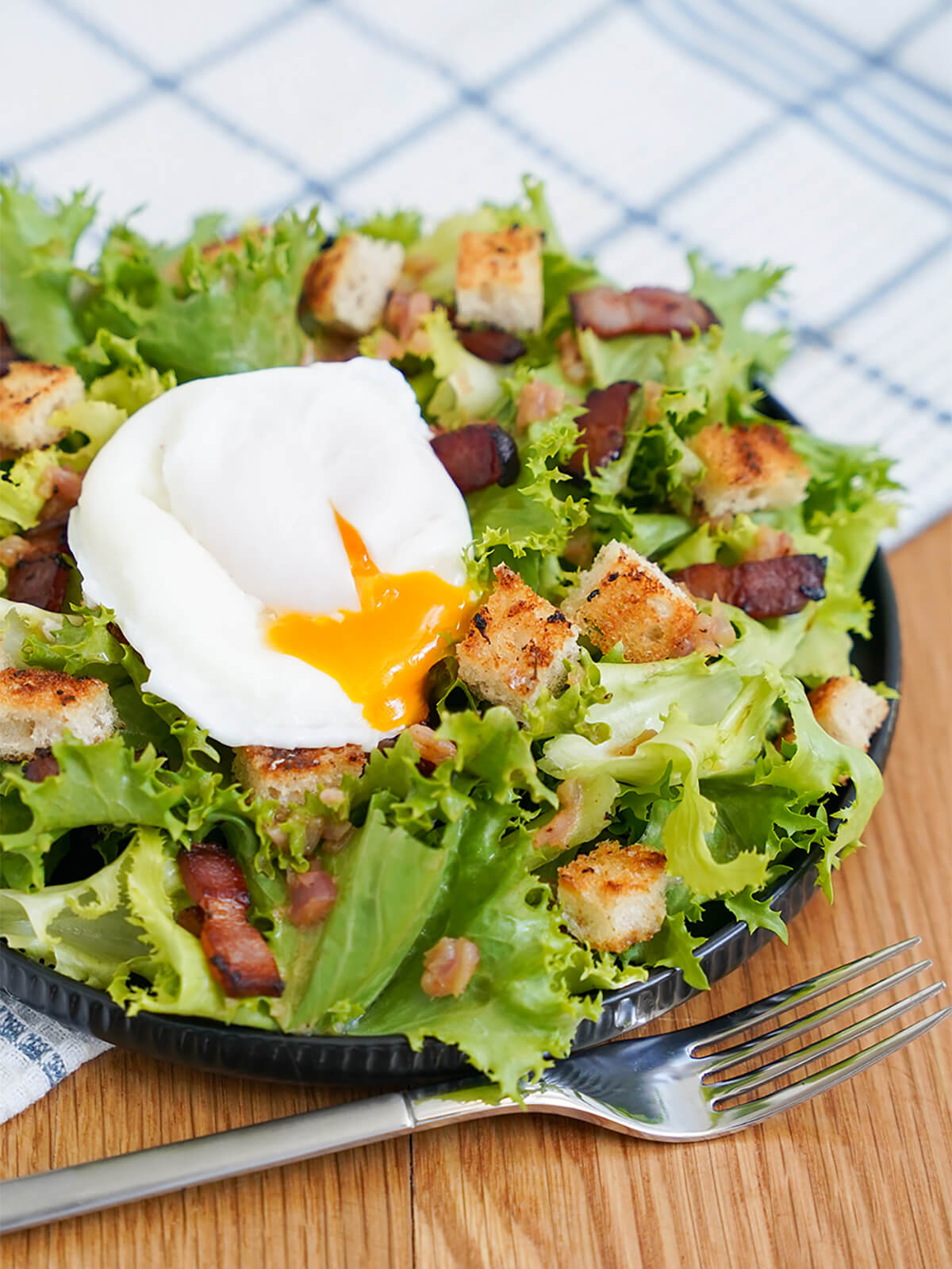side view of plate of salade lyonnaise with fork below and with egg on top with yolk dripping into salad.