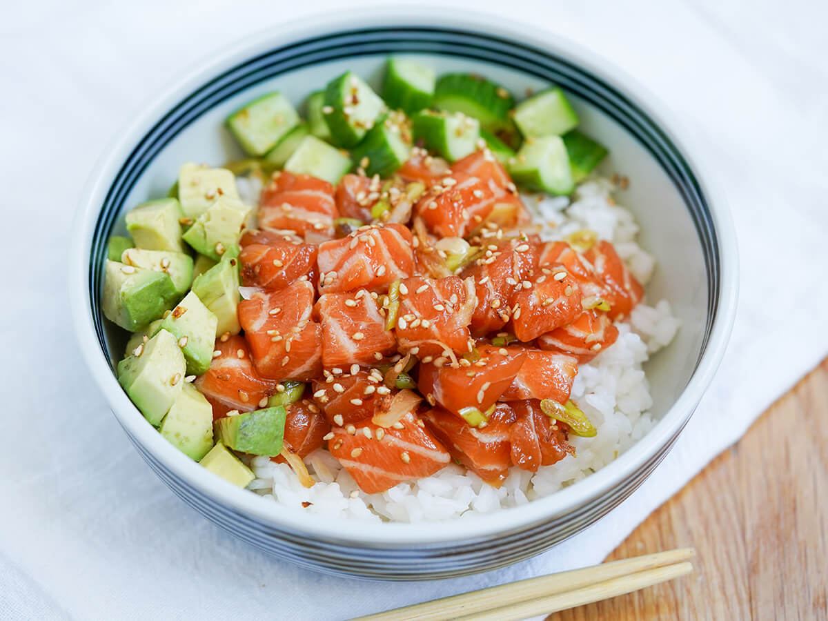 side view of salmon poke bowl with avocado to side and cucumber behind salmon.