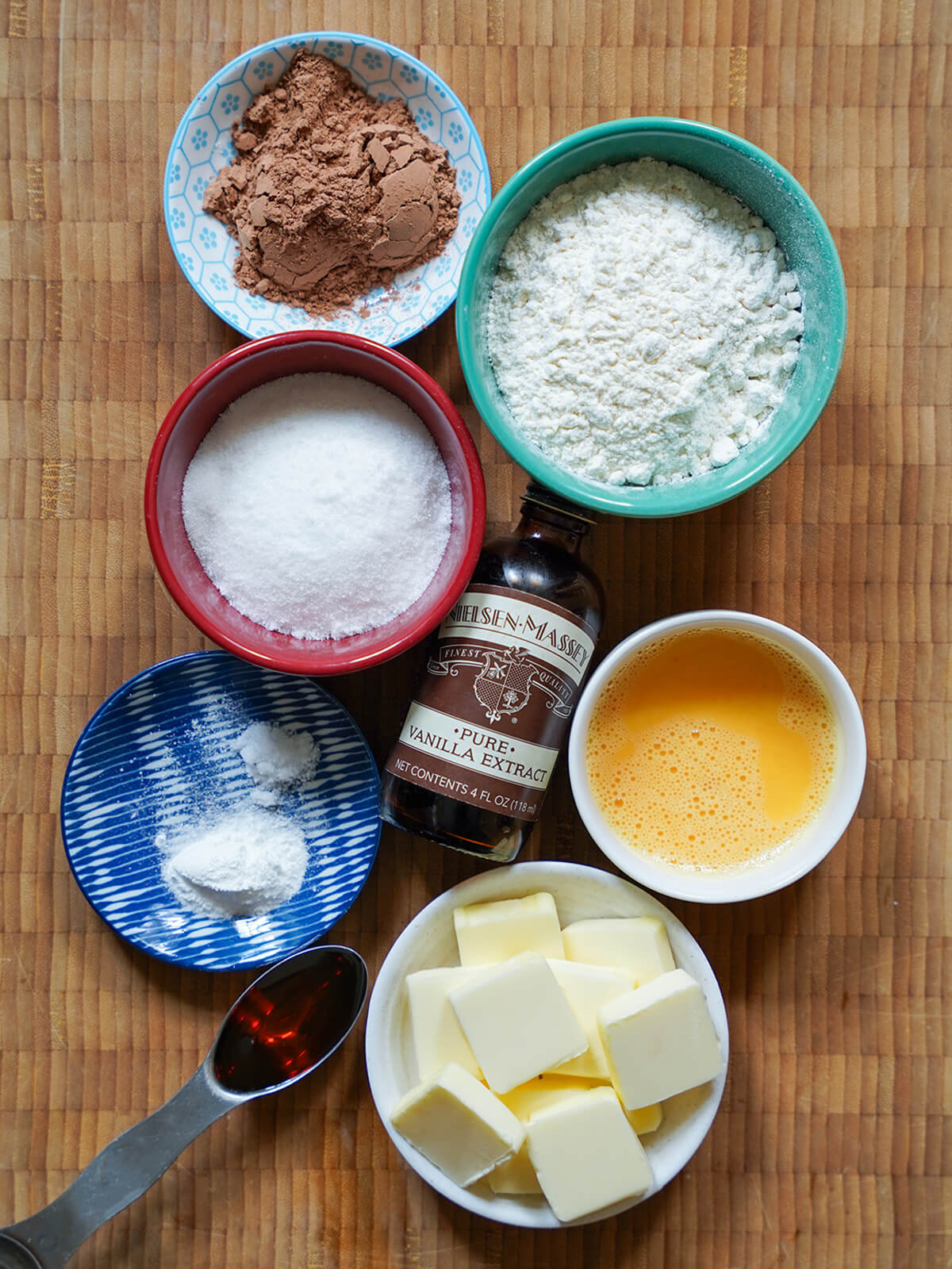 bowls of flour, cocoa powder, sugar, egg, butter with vanilla bottle in middle and spoon of golden syrup to side.