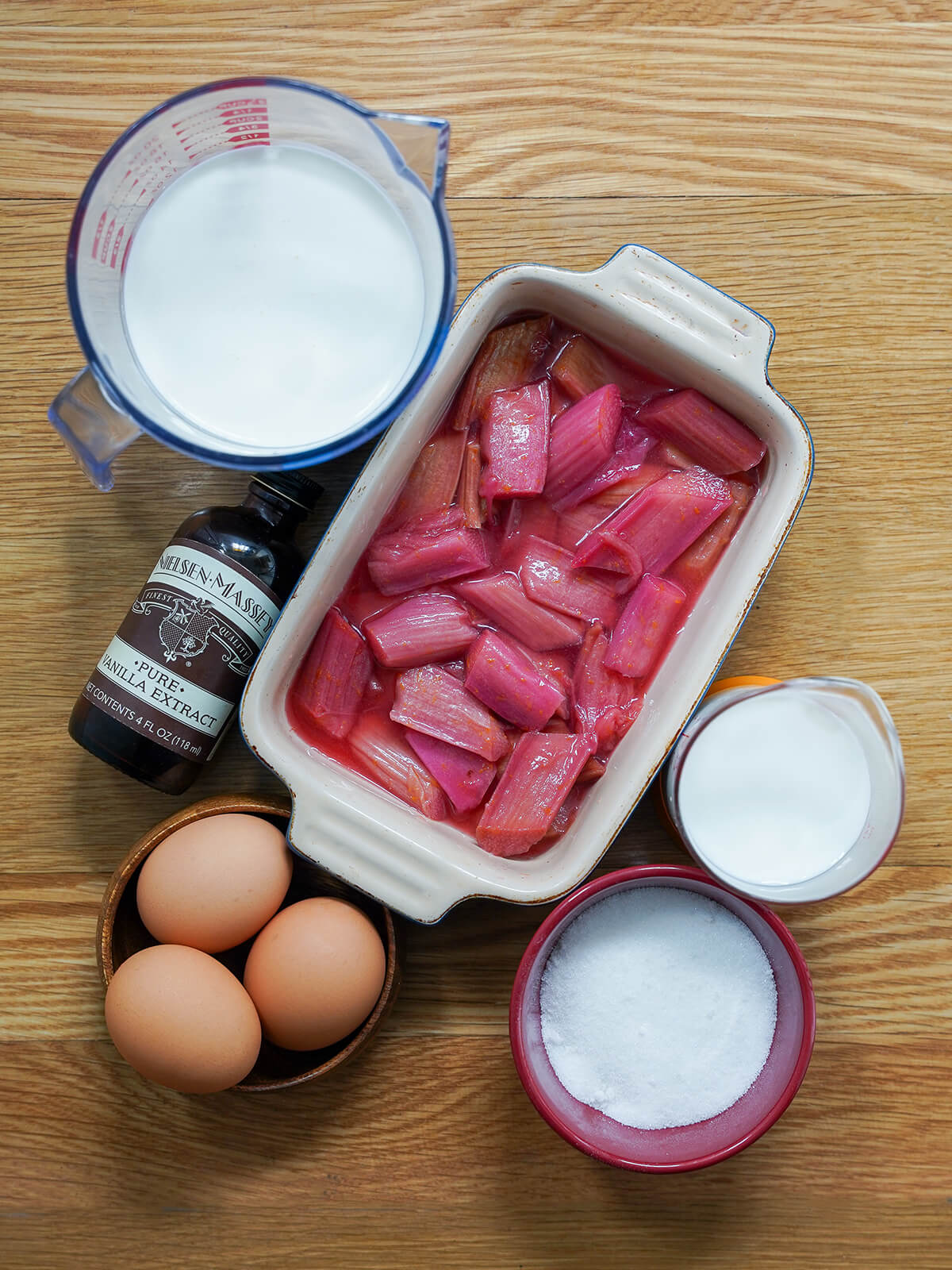 dish with cooked rhubarb surrounded by bowl of sugar, measures with milk and cream and bowl of eggs.