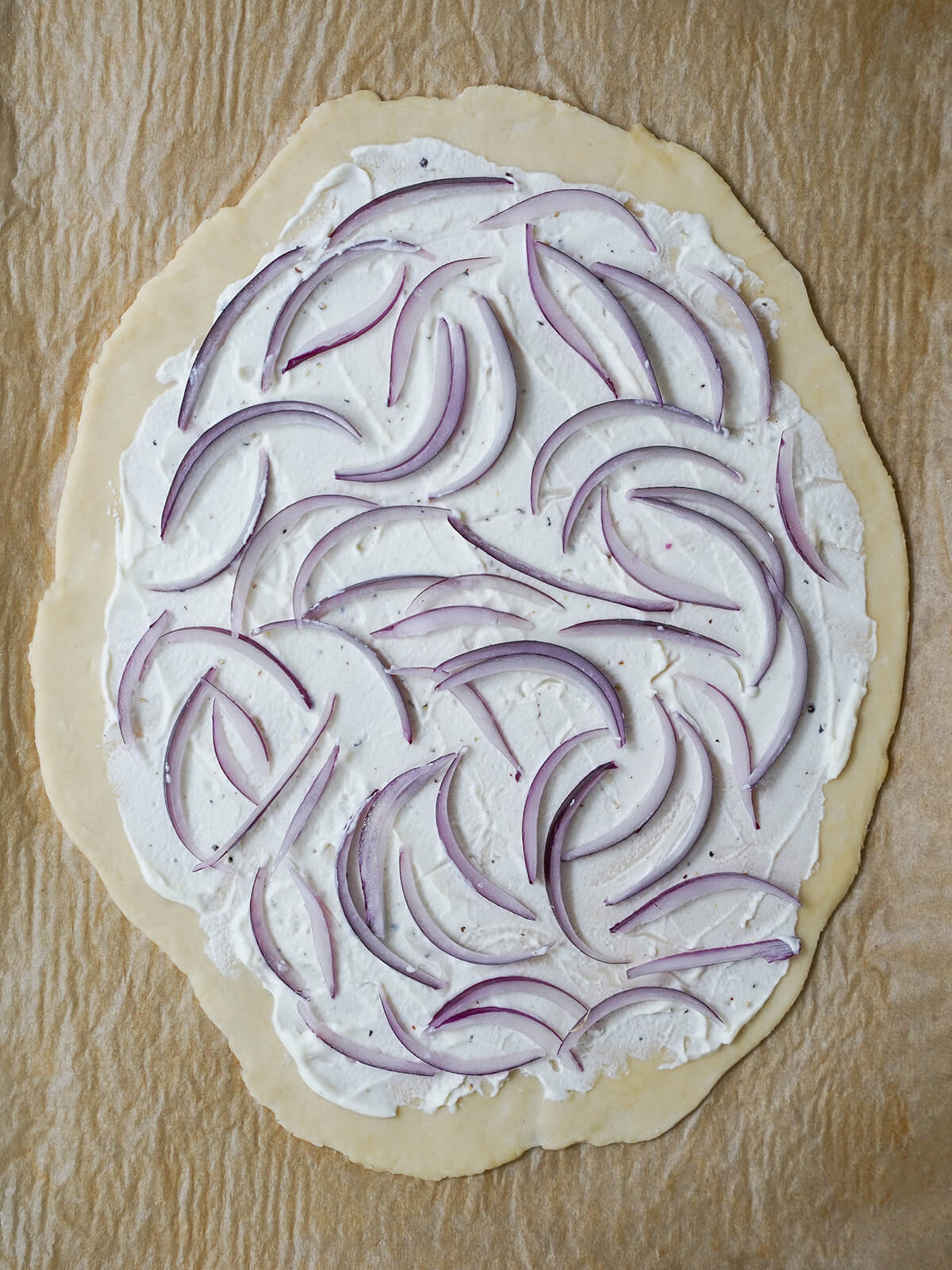 adding creamy layer and onions for smoked salmon pizza dough base with layer of cream spread on top and slices of red onion spread over cream layer.